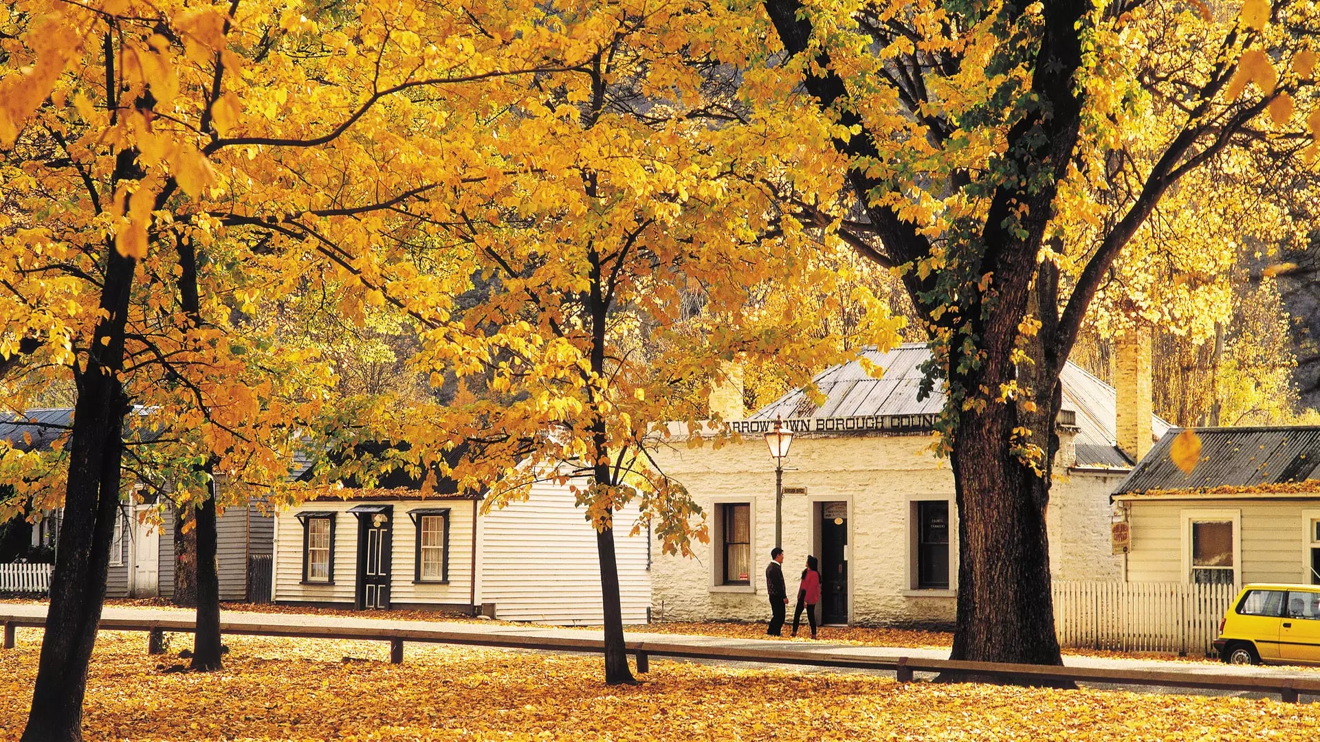 Yellow autumn leaves on trees and ground with historic buildings and two people walking in Arrowtown.
