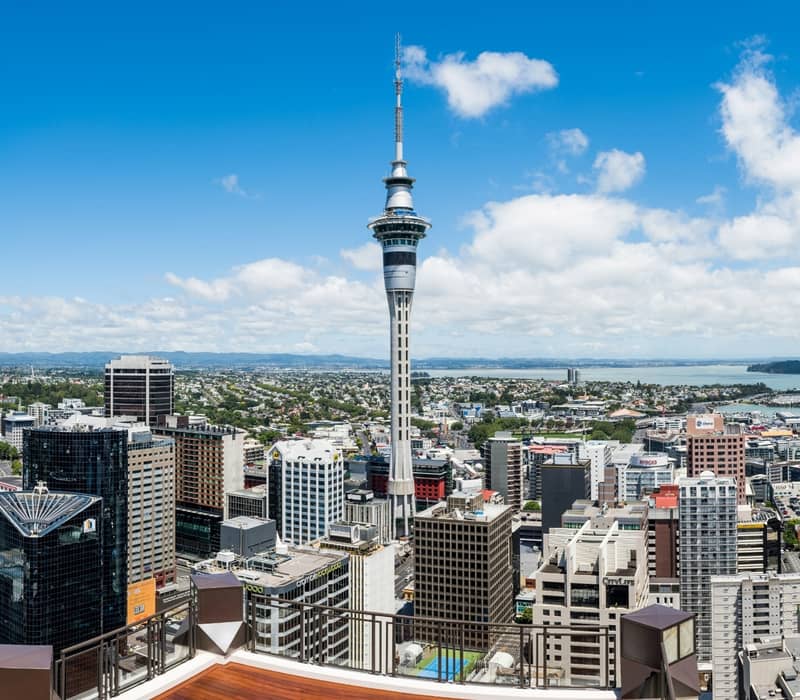 A high-angle view of the Auckland city skyline featuring the iconic Sky Tower under a bright blue sky with fluffy white clouds, with the Waitematā Harbour and suburban hills in the distance.