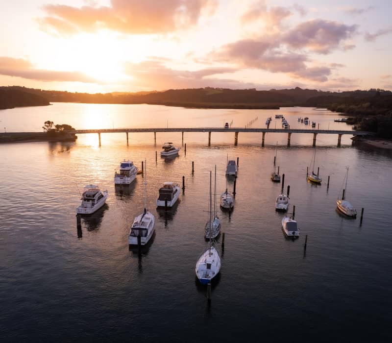 Boats moored in Bay of Islands at sunset with golden light over calm water and a long pier.