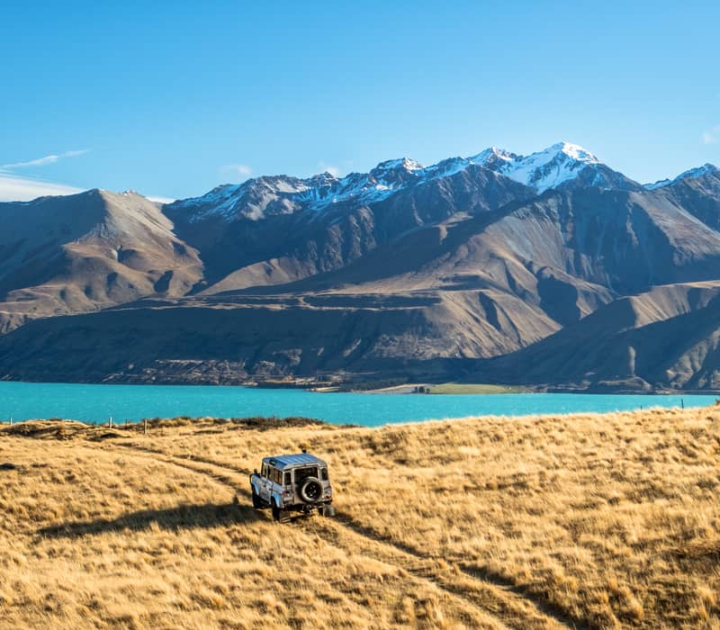 4WD vehicle on grassy plain with lake and snow-covered mountains in background under blue sky