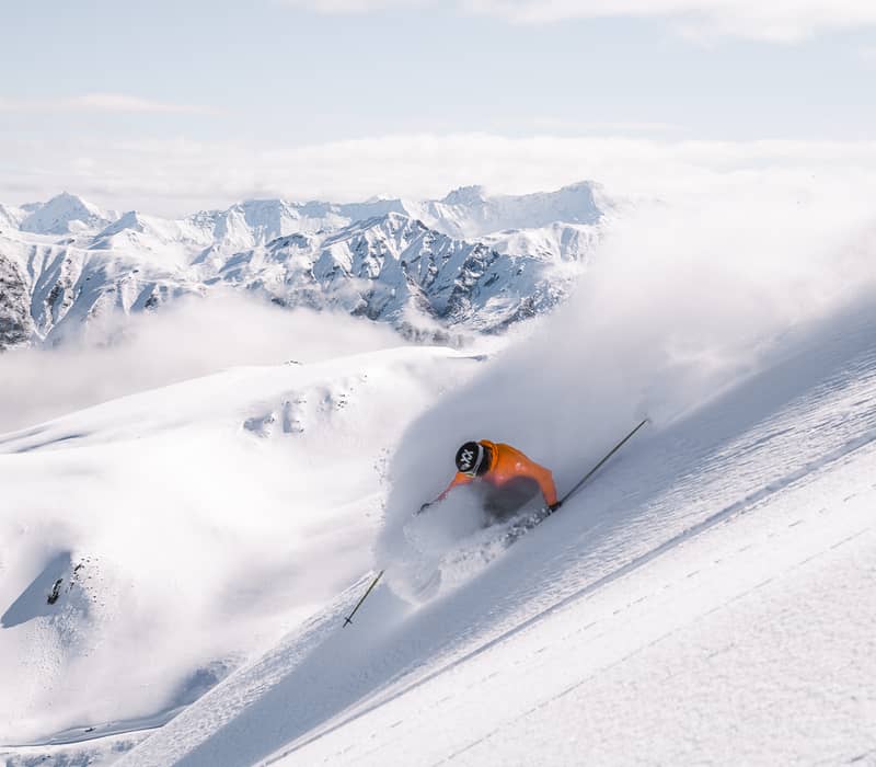 A skier in a bright orange jacket is almost buried in a cloud of deep, white powder as they ski down a steep, ungroomed mountain face in Soho Basin.