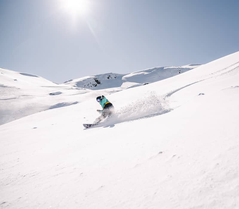 A snowboarder in a teal and yellow jacket slashes through fresh, white powder in the Soho Basin, creating a large "rooster tail" spray of snow.