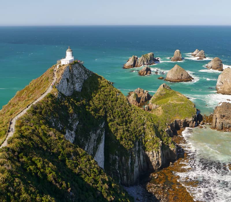 White lighthouse perched on a rocky cliff with a path and jagged rock formations in the blue sea below.