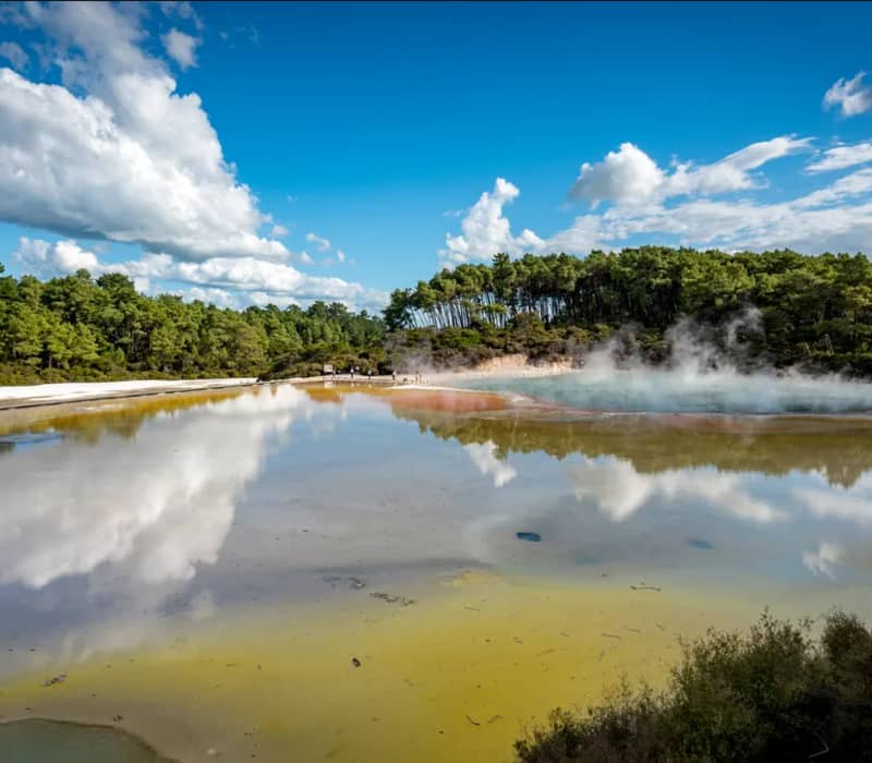 A wide panoramic view of the Champagne Pool in Rotorua, featuring vibrant orange and yellow mineral edges, steaming turquoise water, and a perfect reflection of white fluffy clouds in the still geothermal lake.