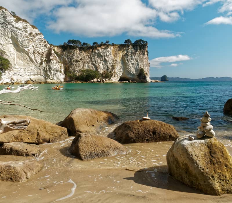 Rocky sandy beach with kayakers on turquoise water and white cliffs in the background under blue sky.