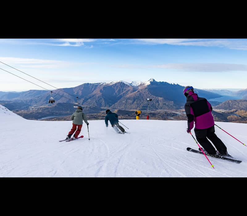 Three skiers in colorful winter gear carve down a wide, groomed trail at Coronet Peak, with chairlifts to the left and a stunning vista of the Queenstown valley and distant mountains in the background.
