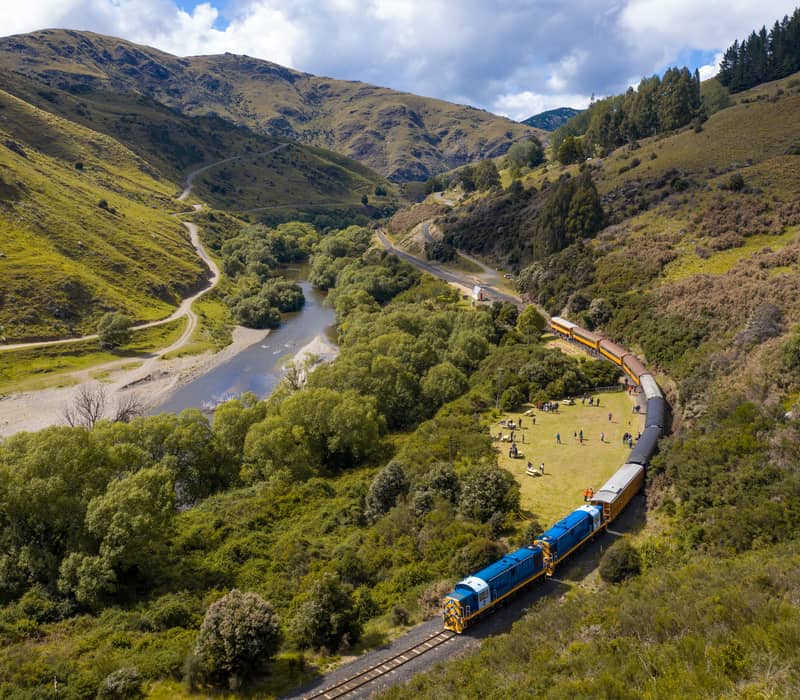 An aerial, high-angle view of a long heritage passenger train with blue and yellow locomotives curving through a deep green valley alongside a winding river, flanked by rugged hills under a blue sky.
