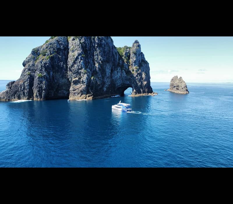 Boat sailing near rocky island with natural arch in blue ocean under clear sky.