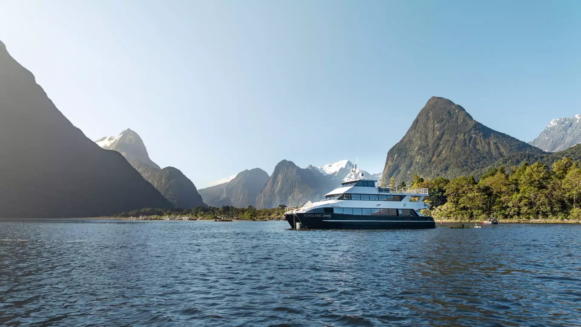 Boat on clear water with forested mountains and blue sky in the background.