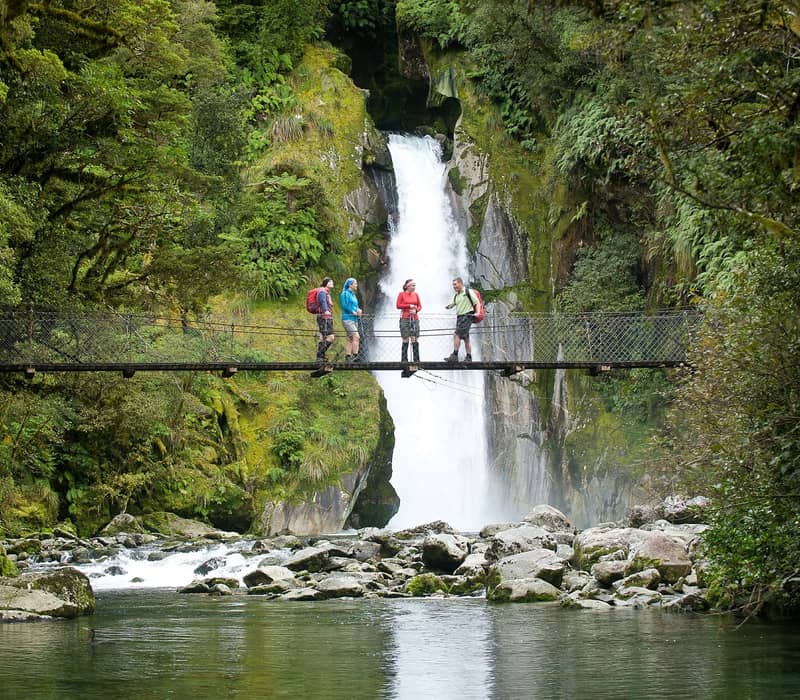 Group of hikers stand on suspension bridge in front of a waterfall in a dense green forest.