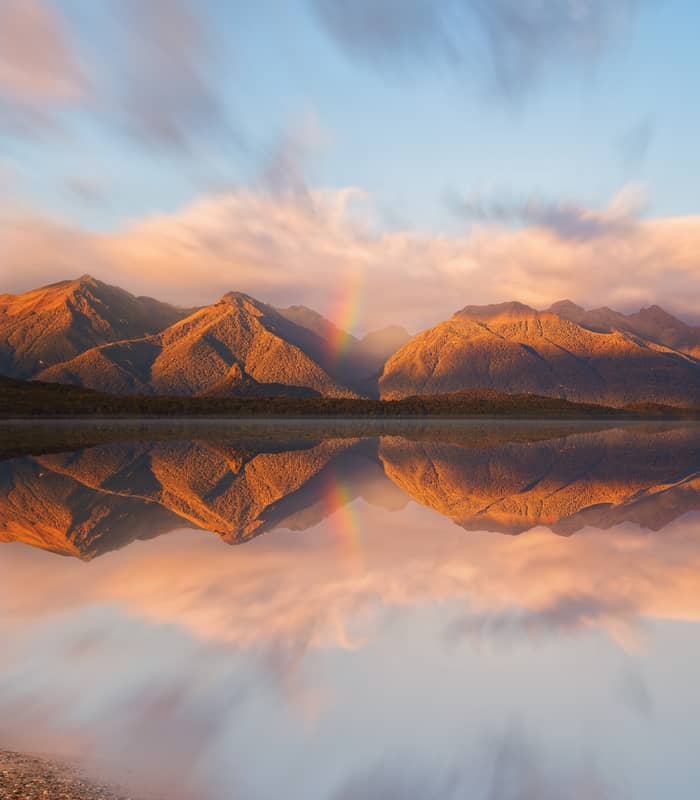 Lake Manapouri reflecting mountains, clouds, and a rainbow at sunset
