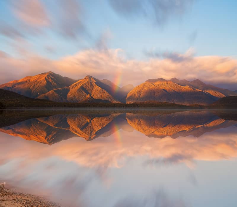 Lake Manapouri reflecting mountains, clouds, and a rainbow at sunset