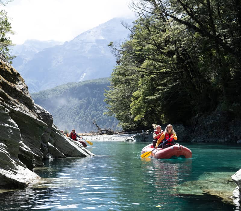 Two rafts float on a clear river between rocky cliffs and green forest with mountains in the background.