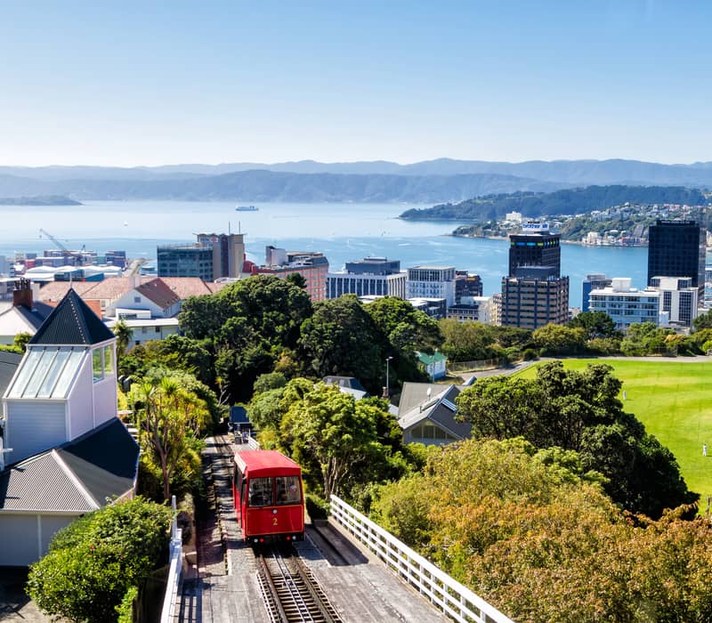 Red funicular tram climbing a green hill with Wellington city, water, and mountains in the background under a blue sky.
