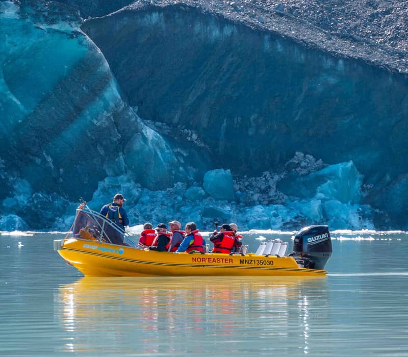Yellow boat carrying tourists with life jackets near blue glacier ice on calm water.