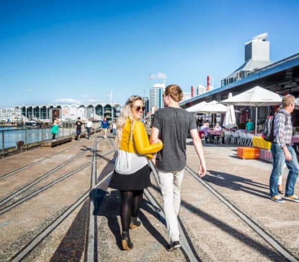Couple walking on waterfront promenade with outdoor dining and buildings under clear blue sky.
