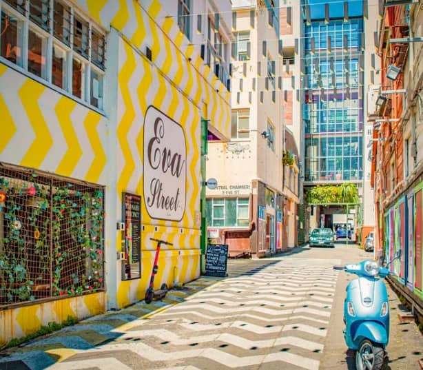 Alley with yellow zigzag walls, blue scooter, and city buildings in background.