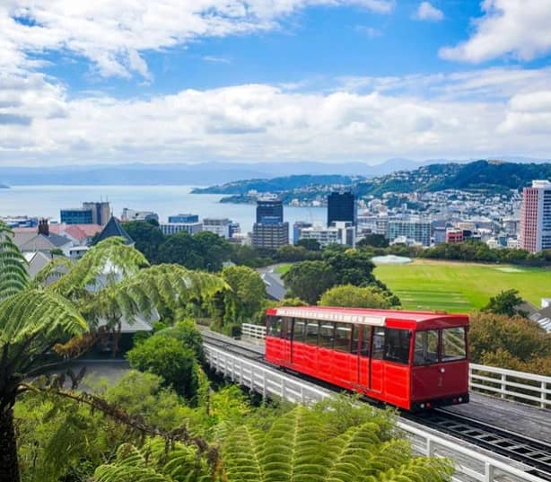 Red cable car on tracks with city buildings, green trees, and water in the background under a cloudy sky.