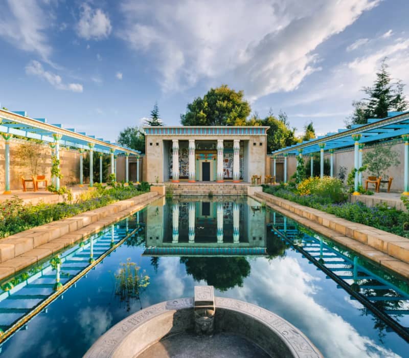 A symmetrical view of a bright blue reflecting pool leading to an ancient Egyptian-style temple with colorful hieroglyphics and painted columns, framed by turquoise pergolas under a bright, cloudy sky.