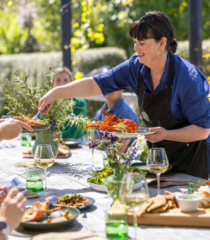 A woman serving lobster outdoors at a table set for lunch with people, glasses, and greenery around.