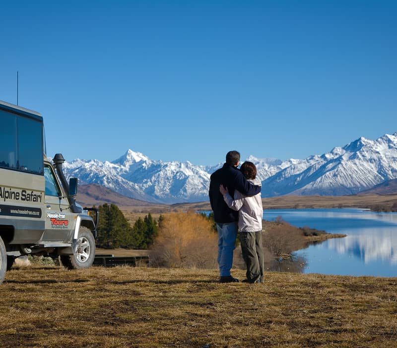 Couple hugging by lake with snow-capped mountains and a safari bus in the background