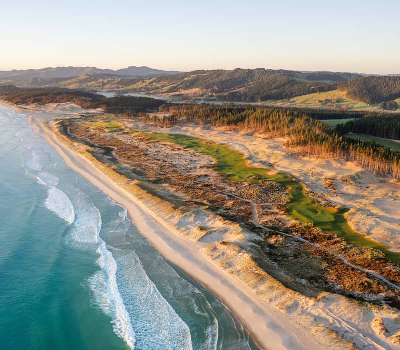 Aerial view of a golf course along a sandy beach coastline with ocean waves and forested hills in the background at sunset.