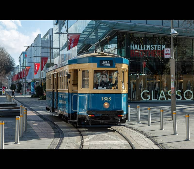 A vintage blue and cream heritage tram, numbered 1888, travels along its tracks in a modern Christchurch pedestrian mall, with large glass-fronted retail stores like Hallenstein Brothers in the background.