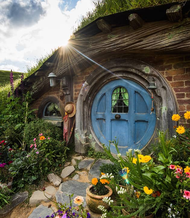Hobbit house with blue round door and colorful flower garden under a partly cloudy sky with sun flare.