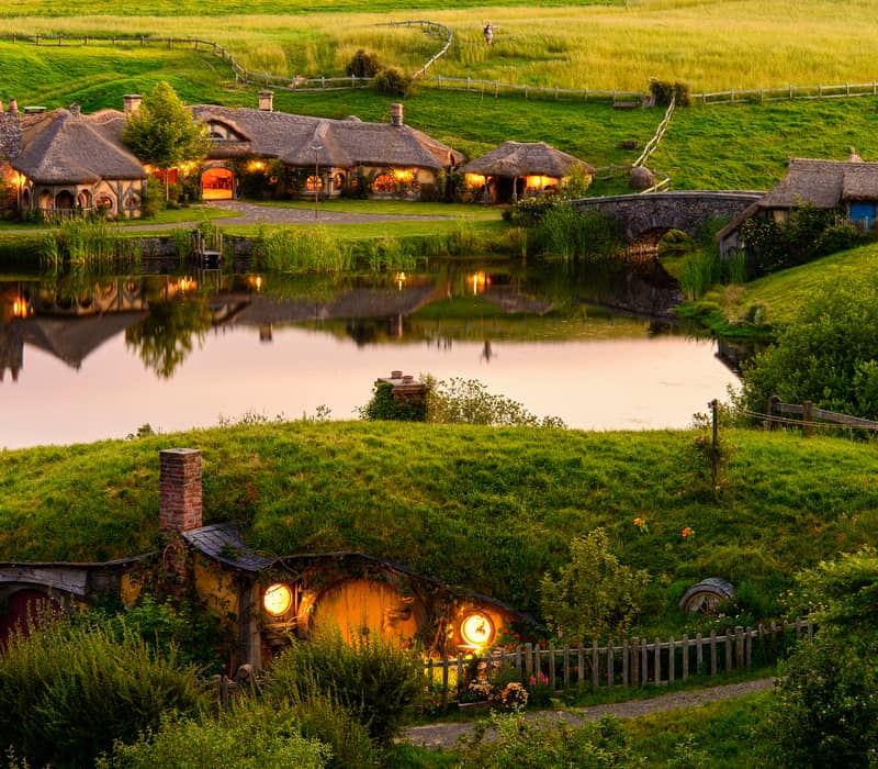 Hobbit houses with glowing windows and thatched roofs by a pond and stone bridge in green countryside at sunset.