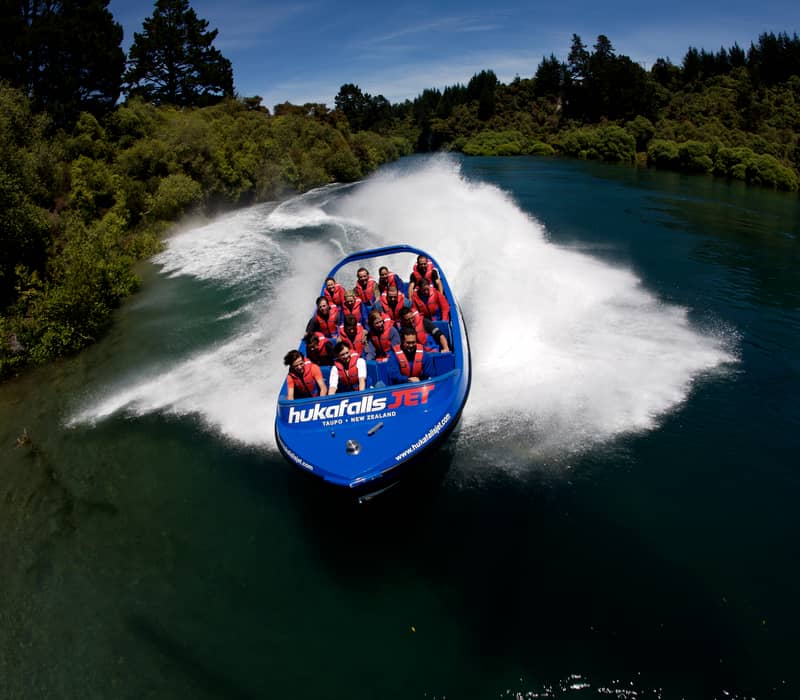 Blue jetboat with people in life jackets making a splash on a river surrounded by trees.