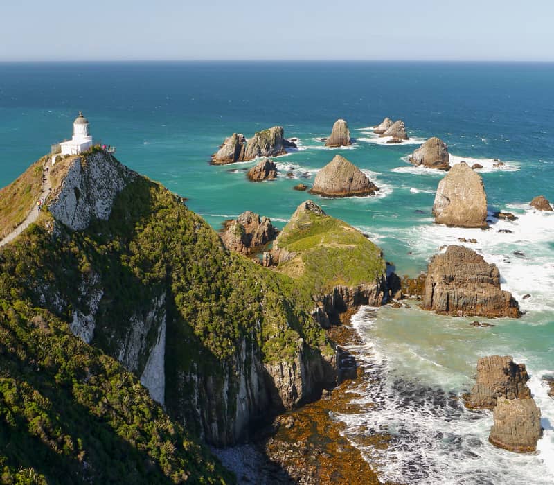 Lighthouse on cliff with rocky ocean formations and turquoise sea below on a sunny day.