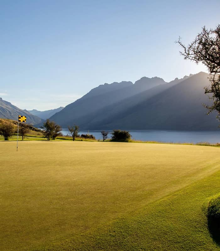 Golf green with flagstick near a lake, mountains in the background, and sunlight streaming through trees.