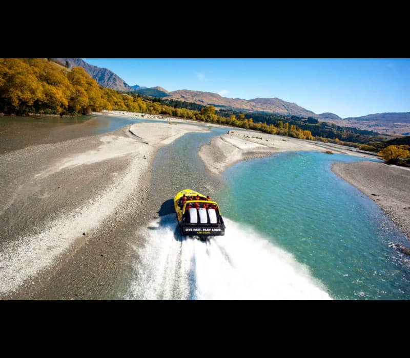 An aerial view of a yellow and black jet boat speeding through a shallow, turquoise braided river, leaving a white wake, surrounded by golden autumn trees and rugged mountains under a clear blue sky.