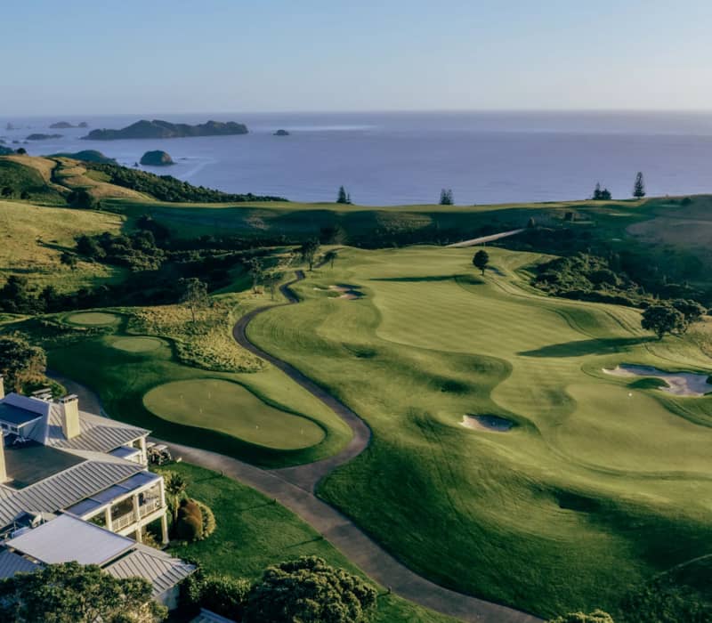 An aerial view of the lush green fairways and bunkers at Kauri Cliffs golf course, overlooking the Pacific Ocean and offshore islands.