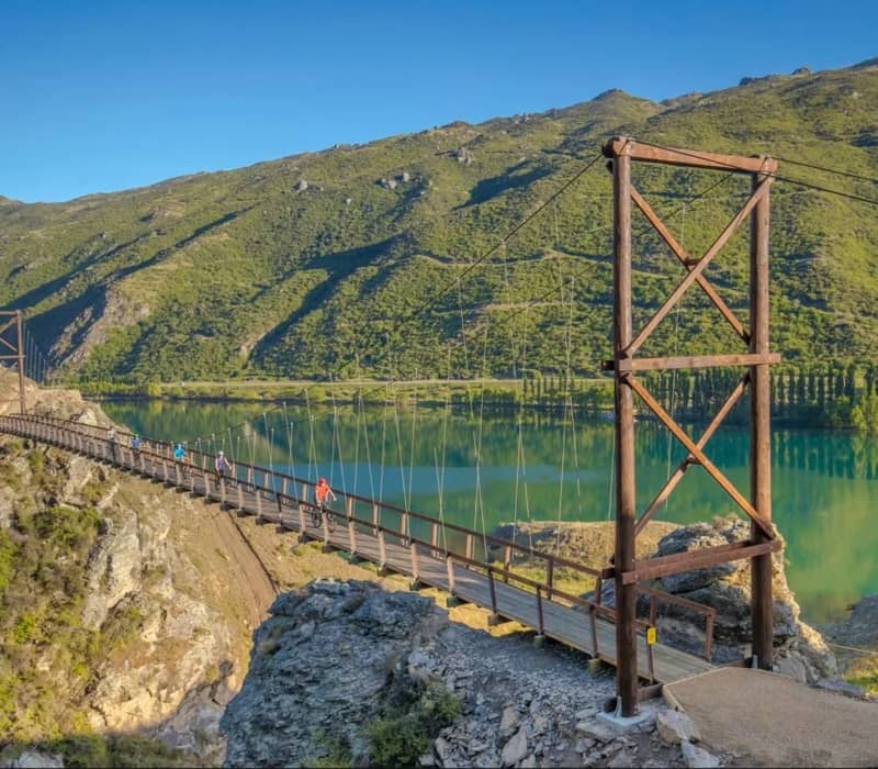 A long wooden suspension bridge with tall timber towers spans a turquoise lake in a rugged, sunlit gorge, with several cyclists riding across it.