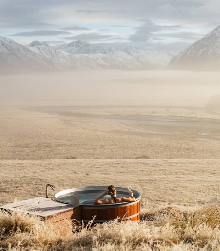 Person in hot tub holding a glass of wine with snowy mountains and misty grassland views.