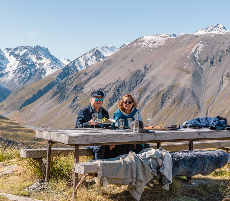 Couple having picnic at wooden table in alpine mountains with snow-capped peaks in background