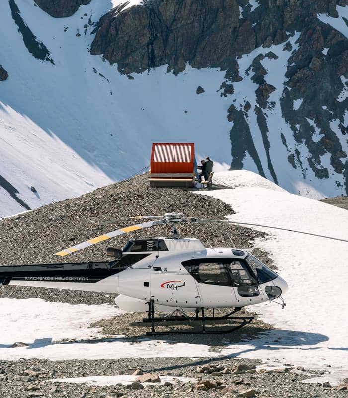 Helicopter on snowy mountain ridge with two people seated near red shelter and rocky peaks behind.