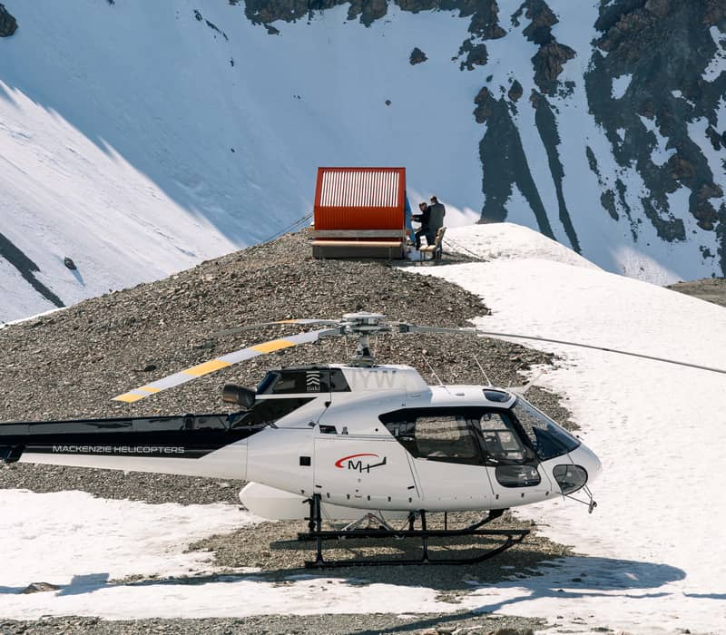 Helicopter on snowy mountain ridge with two people seated near red shelter and rocky peaks behind.