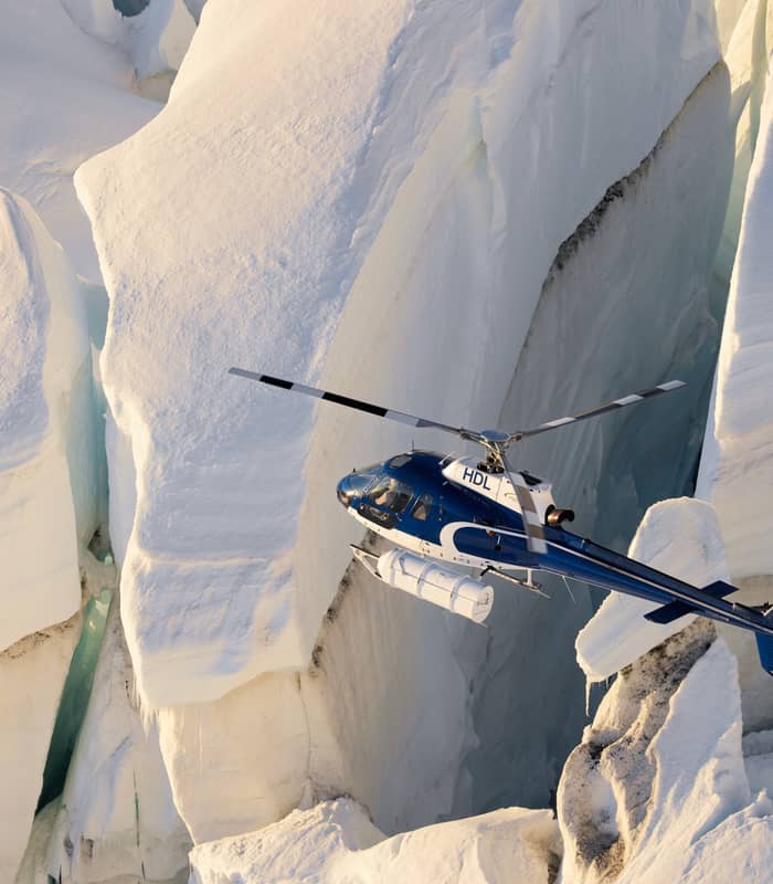Blue helicopter flying near deep crevices in a snow and ice glacier.