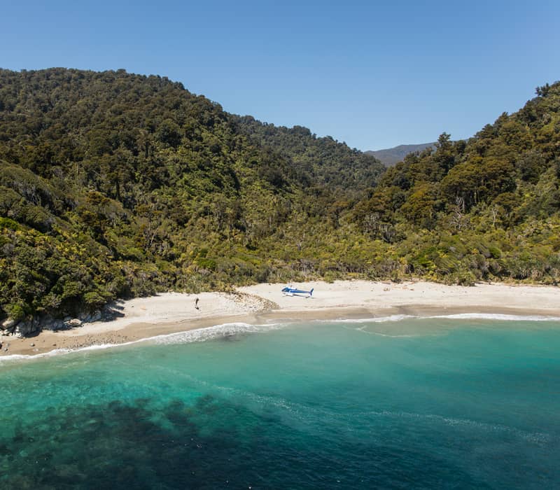 Secluded beach with clear blue water, dense forest behind, and a helicopter on the sand