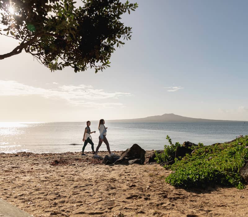 Two women in athletic wear walk along a sandy beach during a bright morning, with a large volcanic island visible across the calm sea and a pōhutukawa tree framing the top of the shot.
