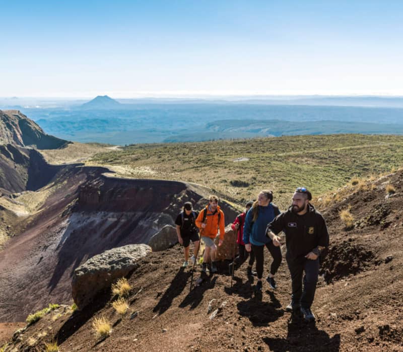 A guided group of hikers walks along the narrow, reddish-brown scree rim of the Mount Tarawera crater under a clear sky, with the vast volcanic landscape and distant peaks of the North Island in the background.
