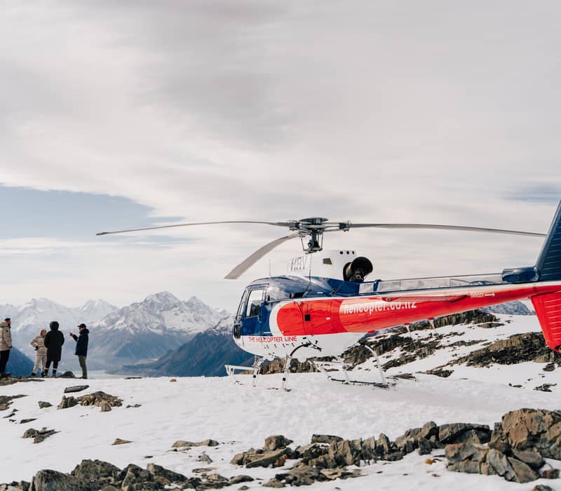 Helicopter on snow with people nearby and snowy mountains in the background under a cloudy sky.