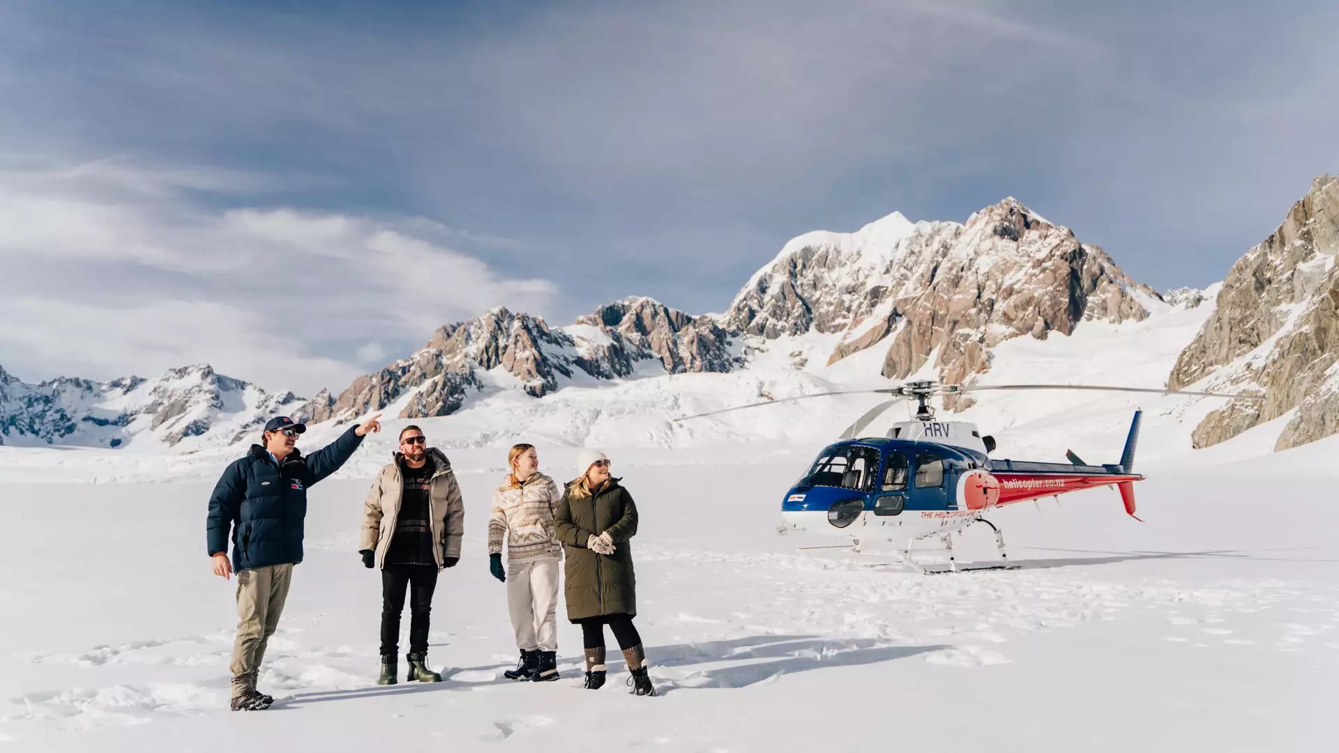 Four people in winter clothes standing on snow near a helicopter with rocky snow-covered mountains behind.