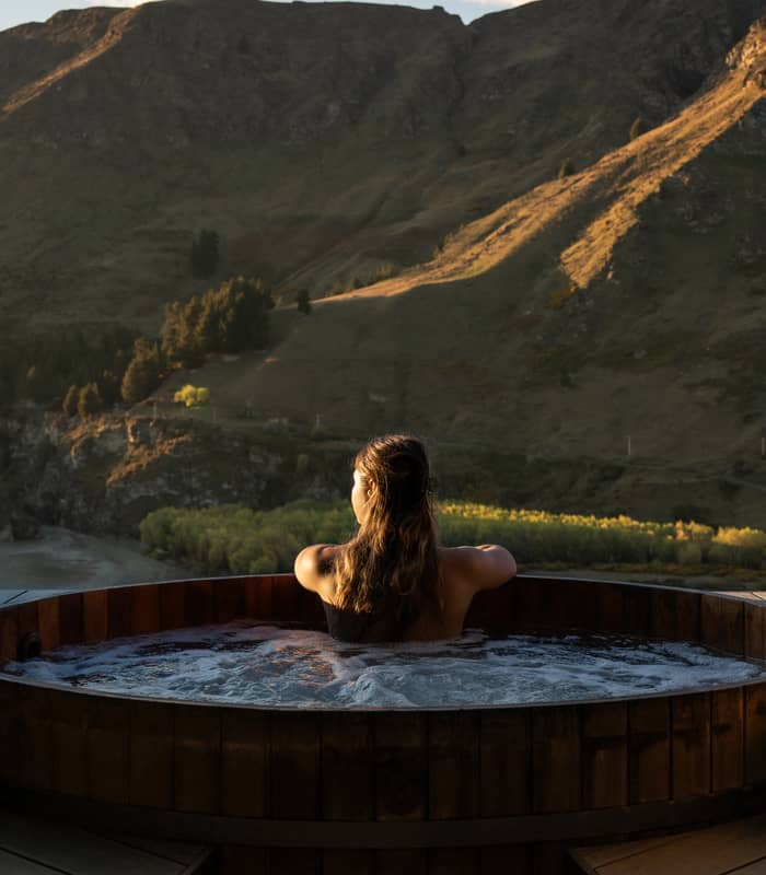 Woman soaking in wooden hot spring tub overlooking mountain landscape at sunset.