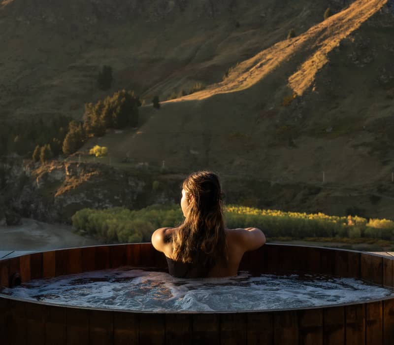 Woman soaking in wooden hot spring tub overlooking mountain landscape at sunset.