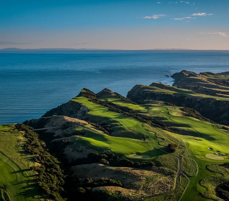 Aerial view of coastal golf course with green fairways and cliffs by the ocean at sunset.