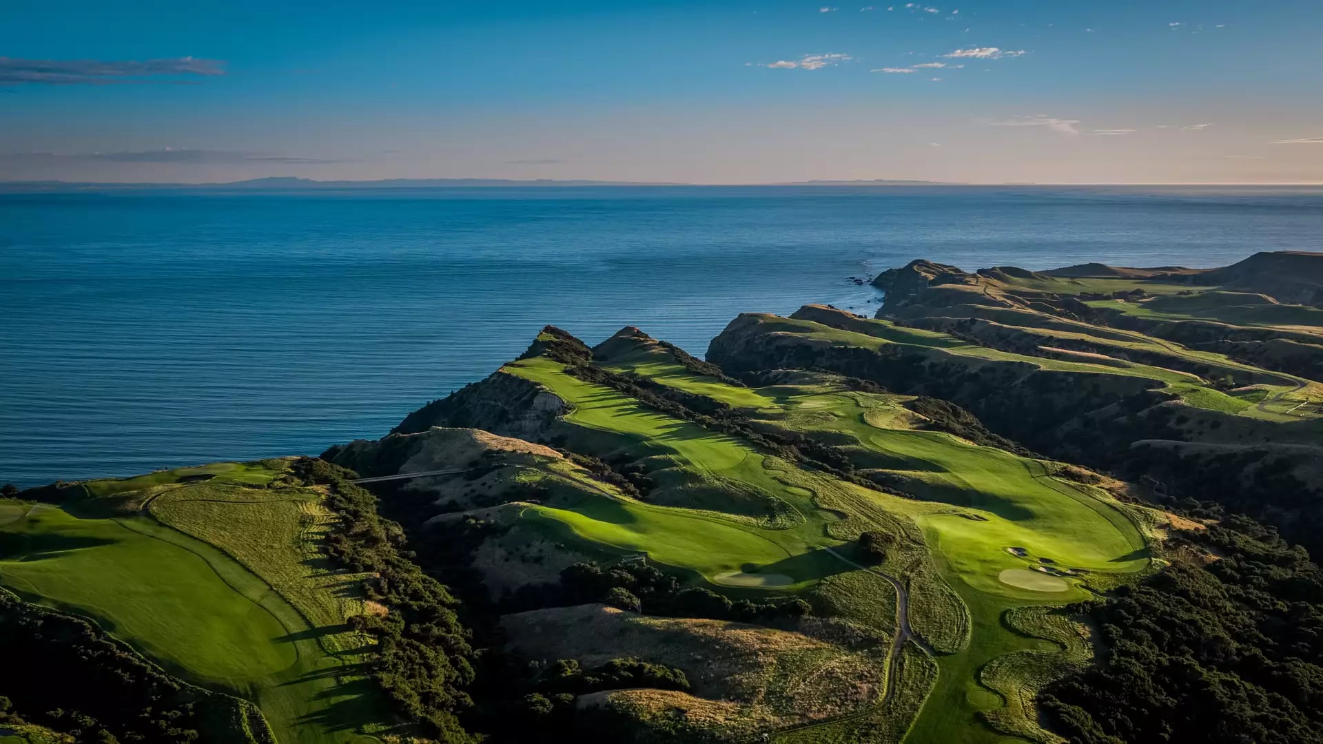 Aerial view of coastal golf course with green fairways and cliffs by the ocean at sunset.