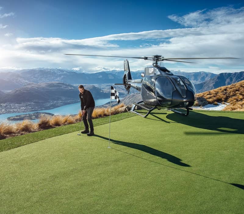 Man golfing on a green next to a helicopter with mountains and a lake behind him.
